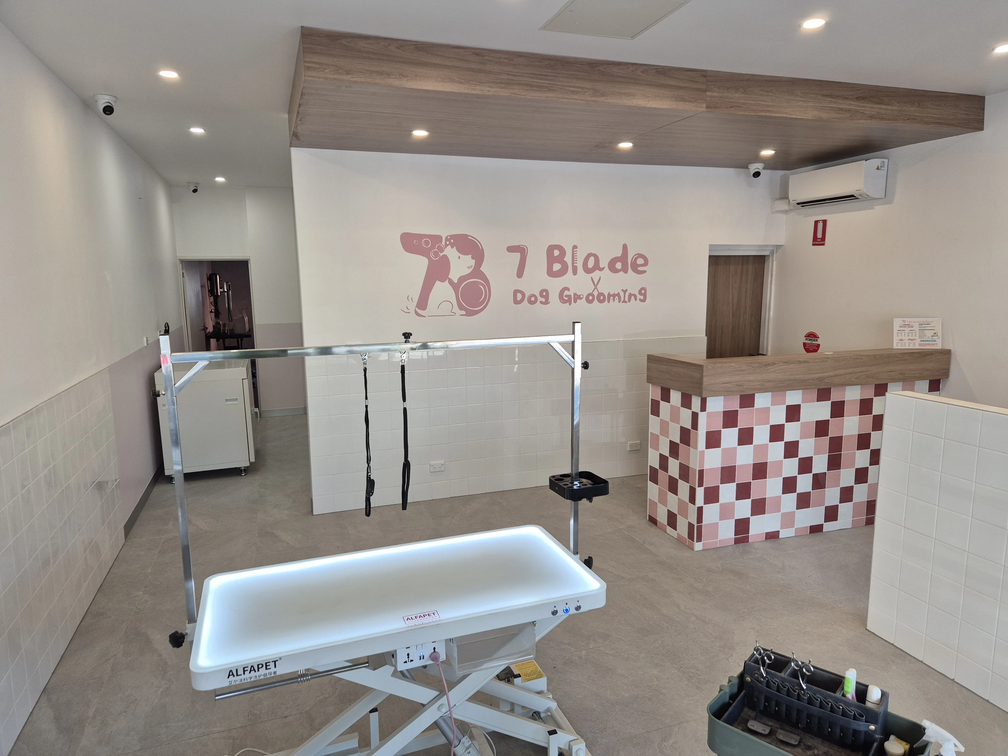 Wide salon interior of 7 Blade Dog Grooming — hydraulic Alfapet grooming table, white subway tiles, pink and burgundy checkerboard reception counter, and the 7 Blade wall mural